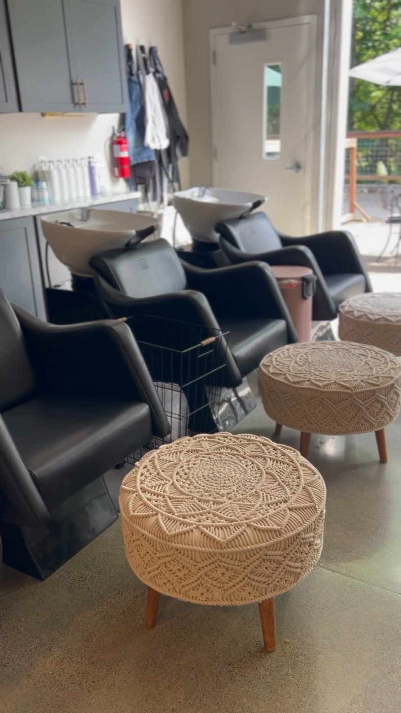 Shampoo row with black backwash chairs and macrame poufs beside open roll-up doors to the patio